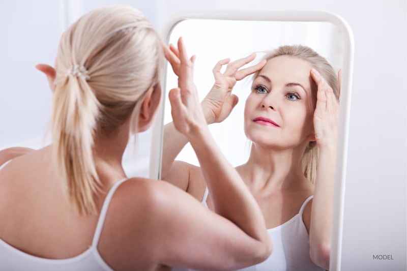 Middle-aged blond woman examining her forehead in the mirror.